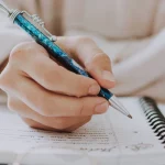 Hand hält blauen Kugelschreiber und schreibt in ein Notizbuch