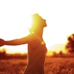 Person steht mit ausgebreiteten Armen in der Abendsonne auf einem Feld