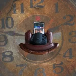 Person sitzt mit Laptop in einem Sessel mit großer Uhr im Hintergrund
